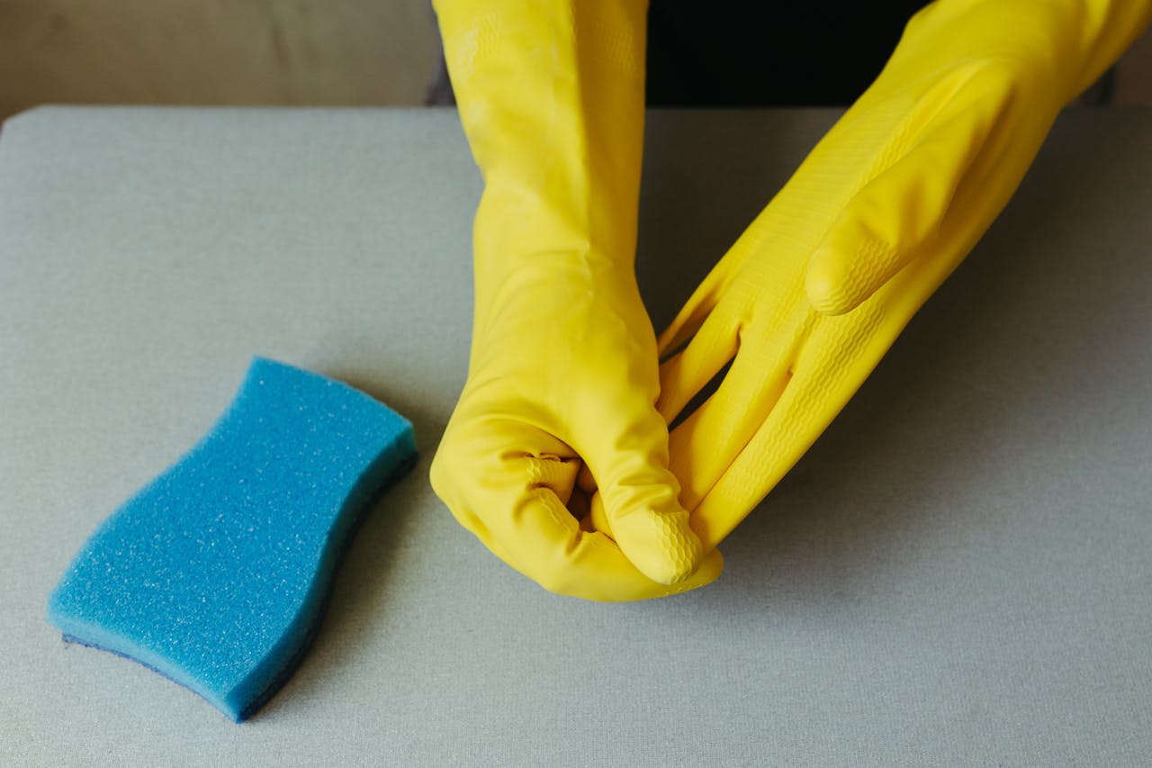services-04 Close-up of hands wearing yellow gloves next to blue sponge on gray surface.