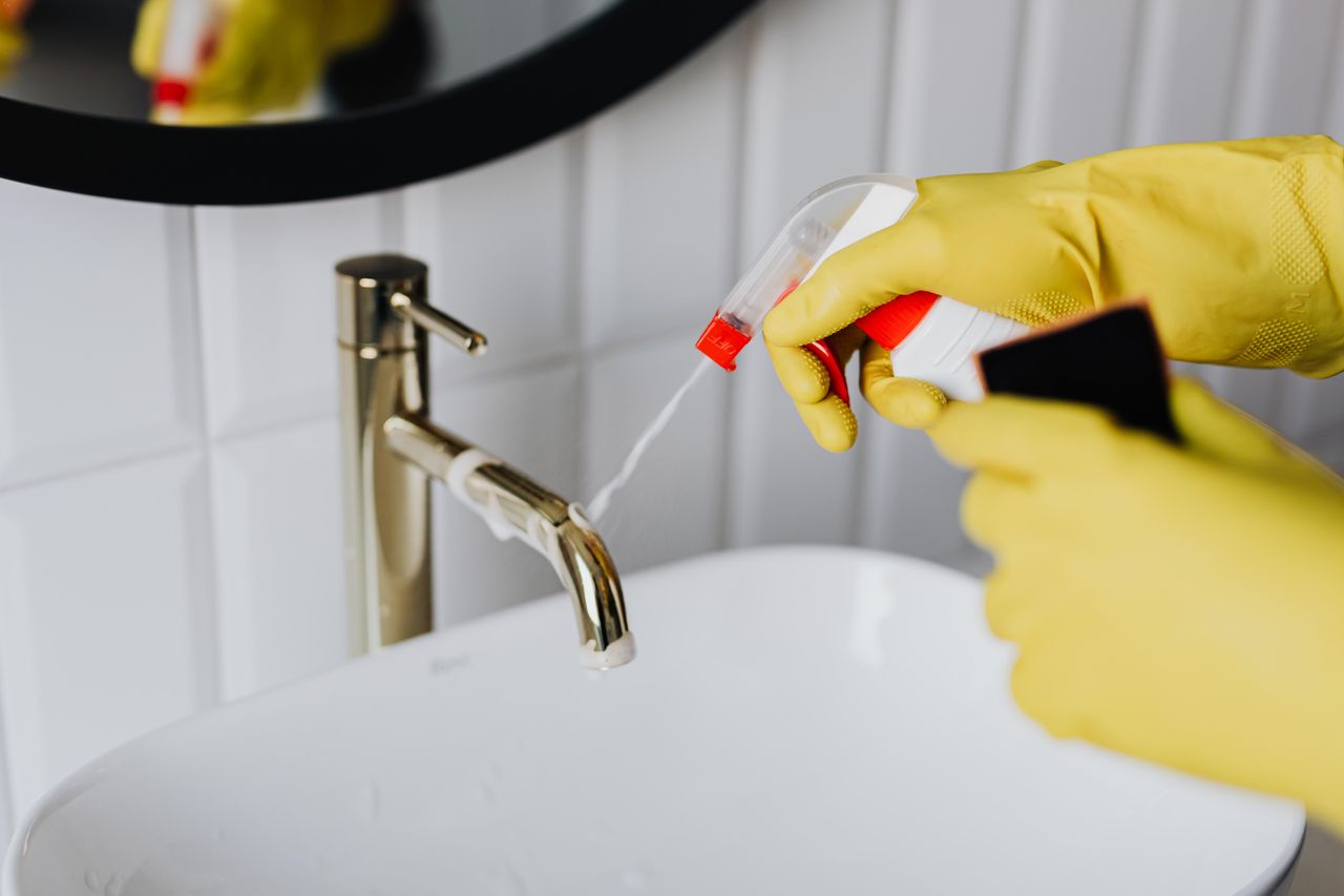 hero-img From above anonymous housekeeper in yellow rubber gloves spraying cleaner to chrome water faucet in bathroom